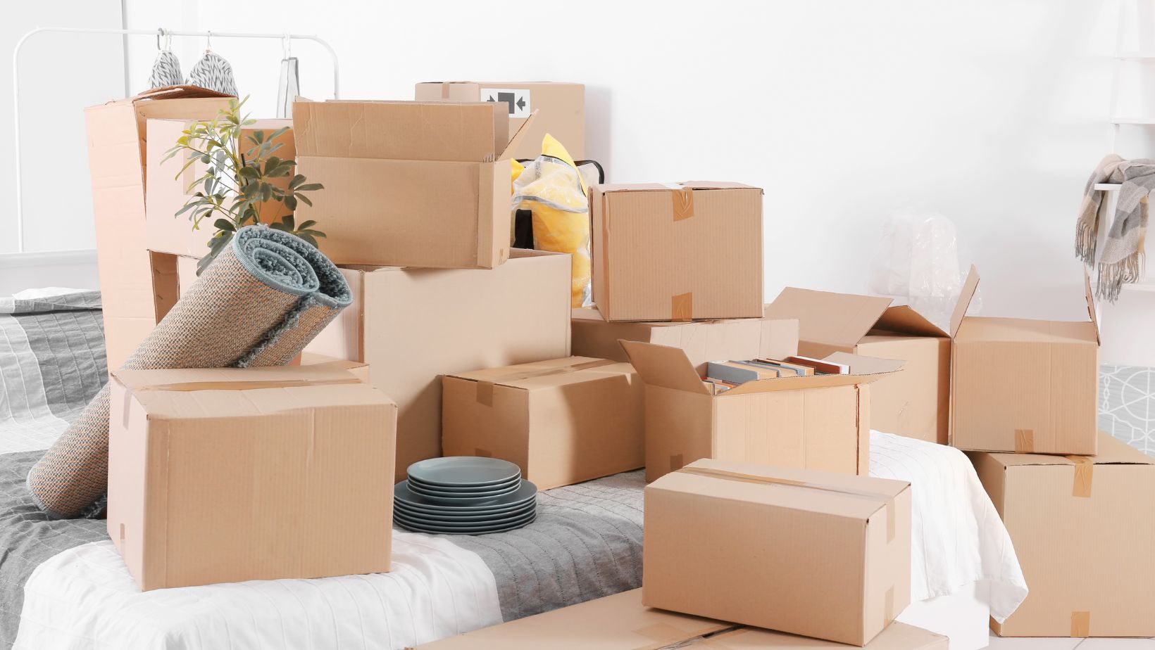 Cardboard moving boxes, household items, books, dishes, and a rolled rug packed in a bedroom during a move.
